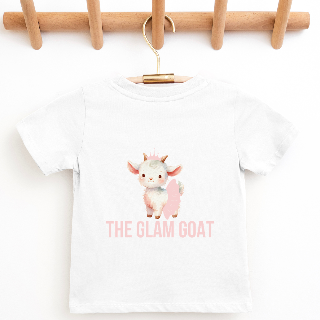 A white short-sleeve baby t-shirt with a cute cartoon goat graphic and the text 'THE GLAM GOAT' in pink, hanging on a wooden hanger from a wooden peg rack. Six pastel tutus are lined up along the bottom of the image.