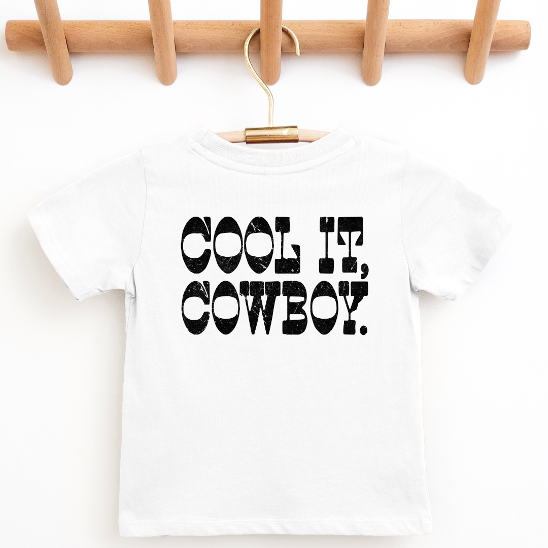 A white short-sleeve baby t-shirt with 'COOL IT, COWBOY.' printed in black distressed letters, hanging on a wooden hanger from a wooden peg rack.