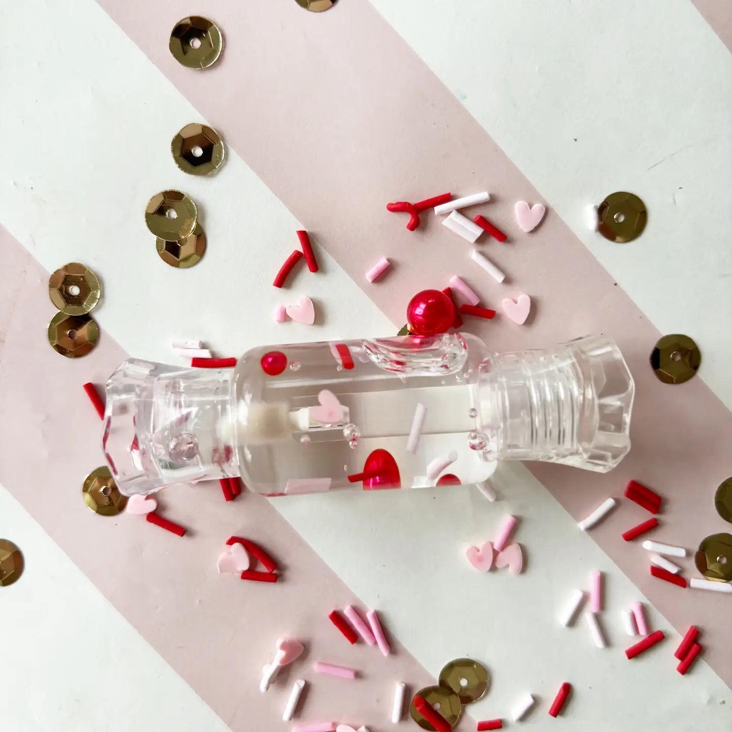 Clear plastic bottle with red cap surrounded by colorful confetti on a pink and white striped background