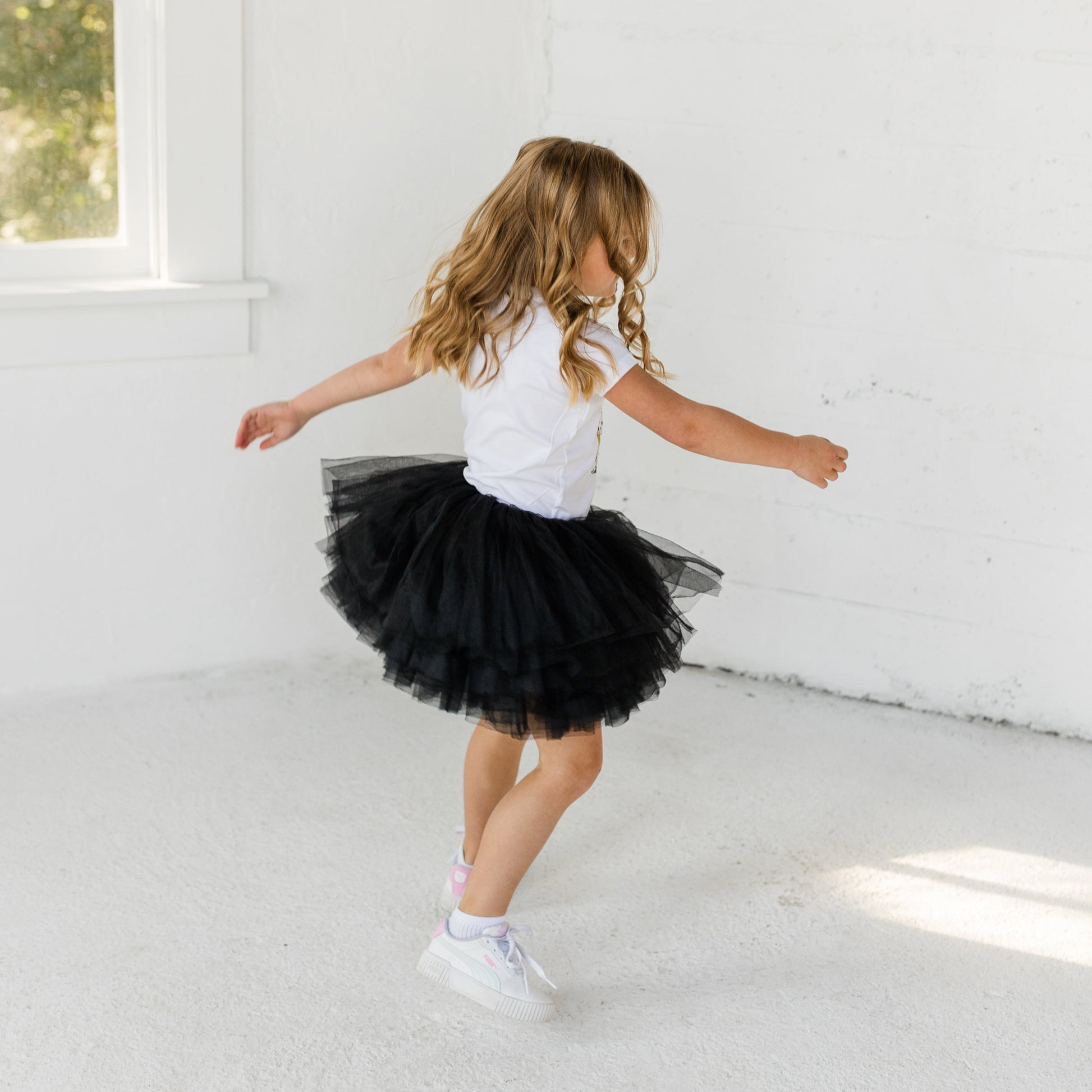 Young girl in a black tutu and white top dancing in a bright room with large windows.