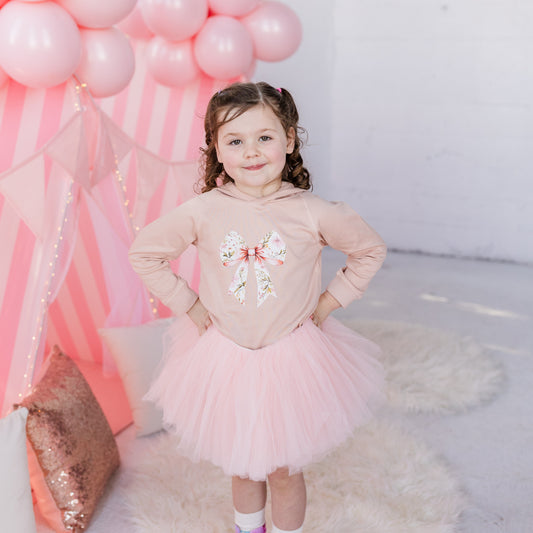 Toddler girl wearing a ballet pink organic cotton hoodie with a floral bow design, styled with a pink tutu, standing in front of pink balloons.