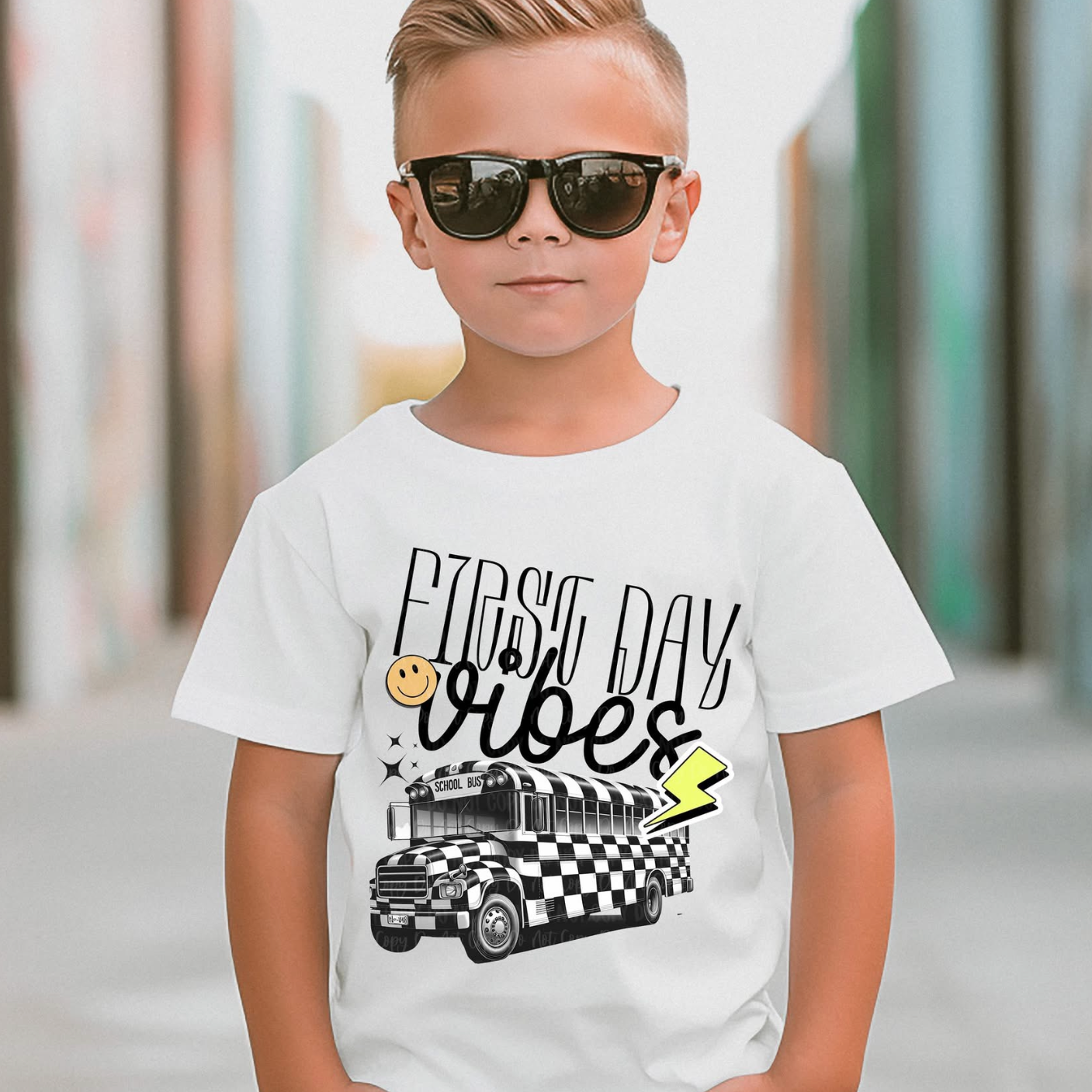 “First Day Vibes” graphic t-shirt with a black-and-white checkerboard school bus, lightning bolt, and smiley face, standing outdoors with sunglasses and a confident look.