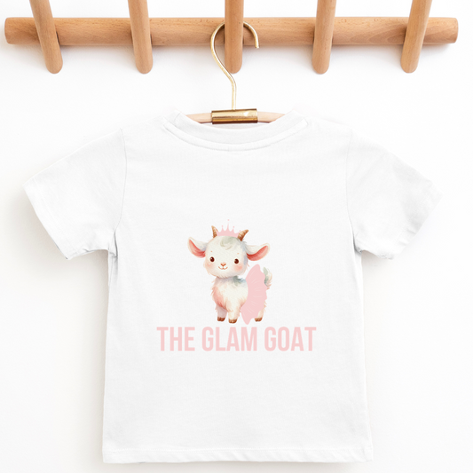 A white short-sleeve baby t-shirt with a cute cartoon goat graphic and the text 'THE GLAM GOAT' in pink, hanging on a wooden hanger from a wooden peg rack. Six pastel tutus are lined up along the bottom of the image.