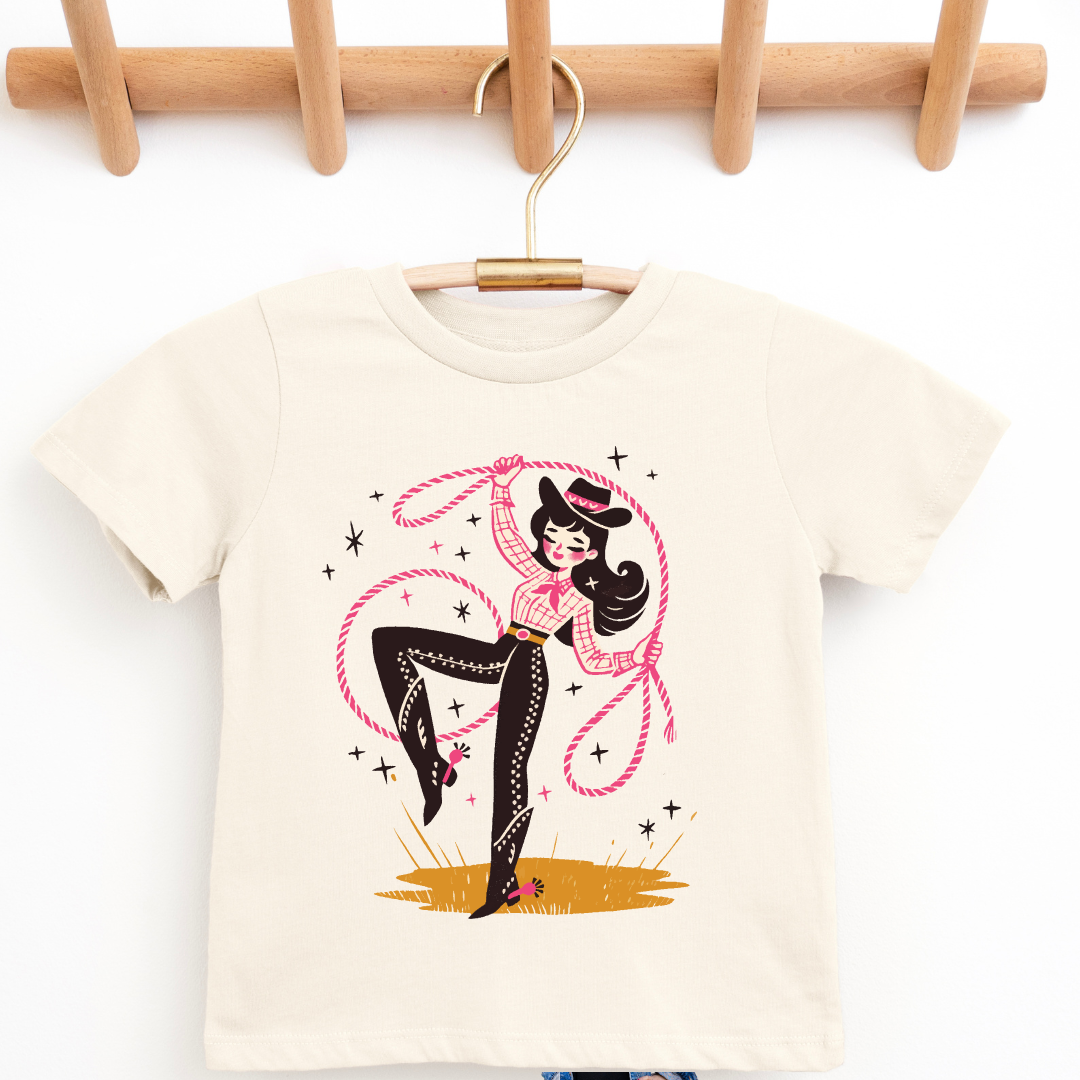 A cream-colored short-sleeve baby t-shirt hanging on a wooden hanger from a wooden peg rack. The shirt features a retro graphic of a pin-up girl with a lasso.