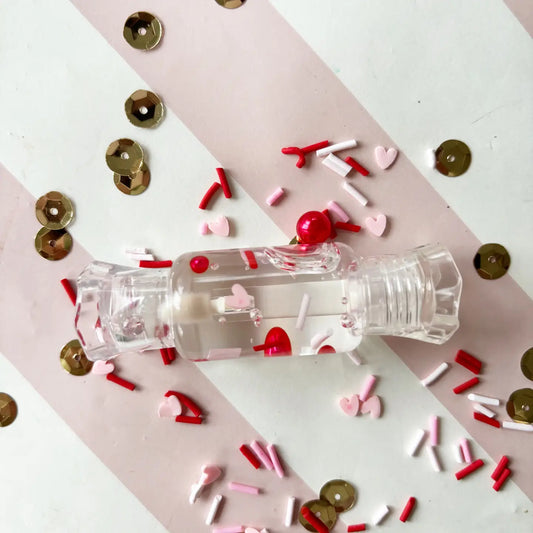 Clear plastic bottle with red cap surrounded by colorful confetti on a pink and white striped background
