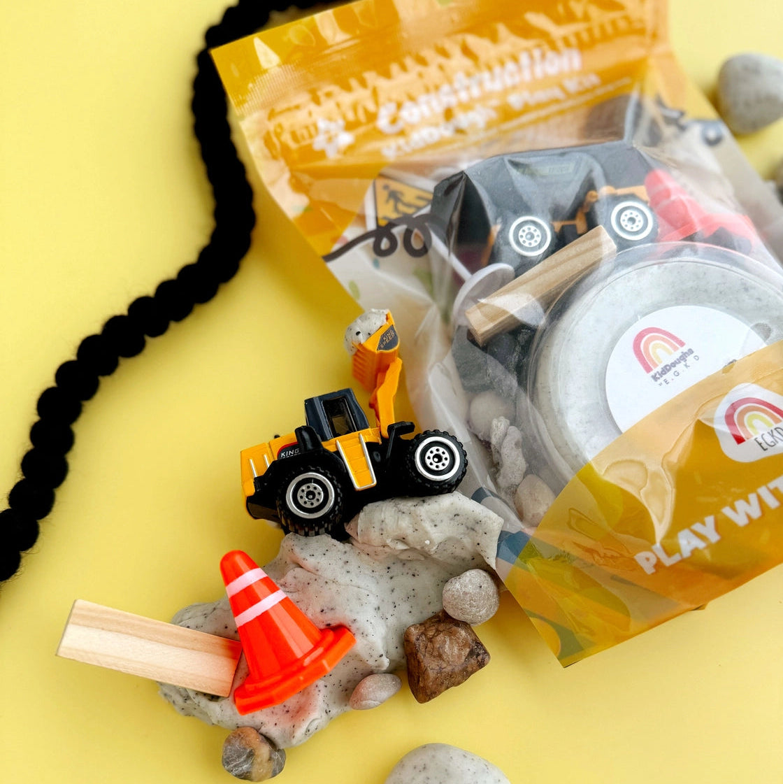 Construction-themed toys and accessories on a yellow background with a 'Play Well' bag.