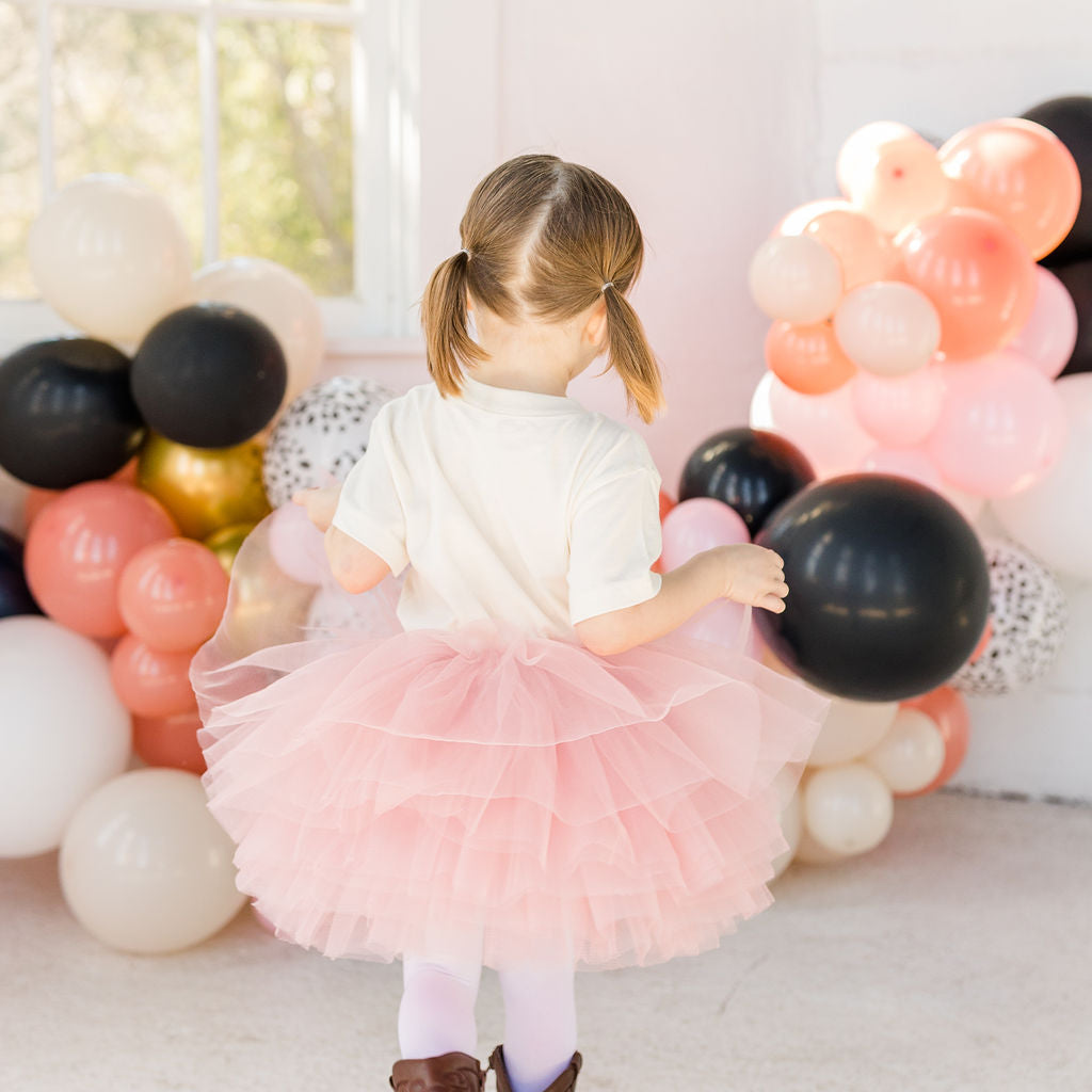 Child in a pink tutu skirt standing among colorful balloons indoors.
