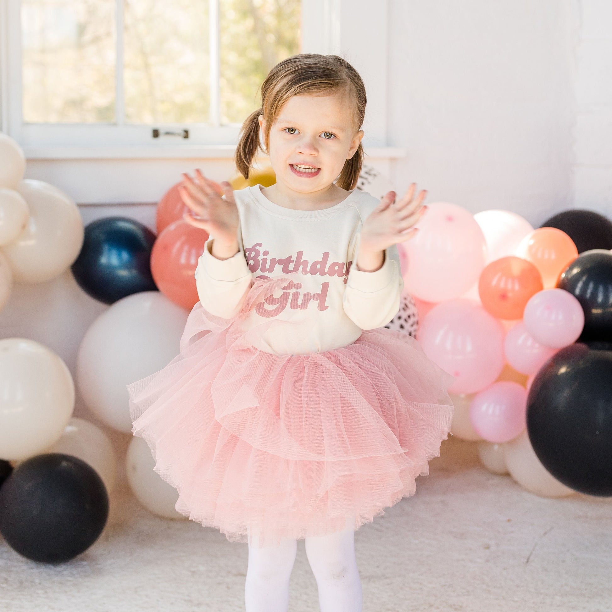 Child in a pink tutu and birthday shirt surrounded by balloons indoors.