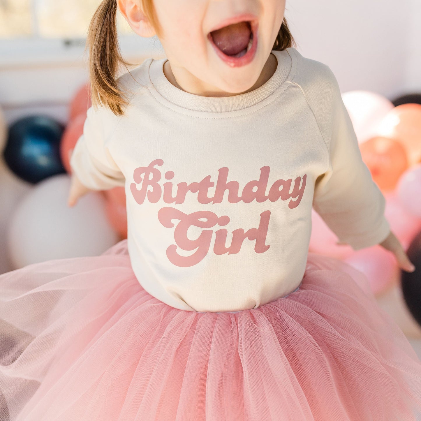 Child wearing a 'Birthday Girl' shirt with balloons in the background