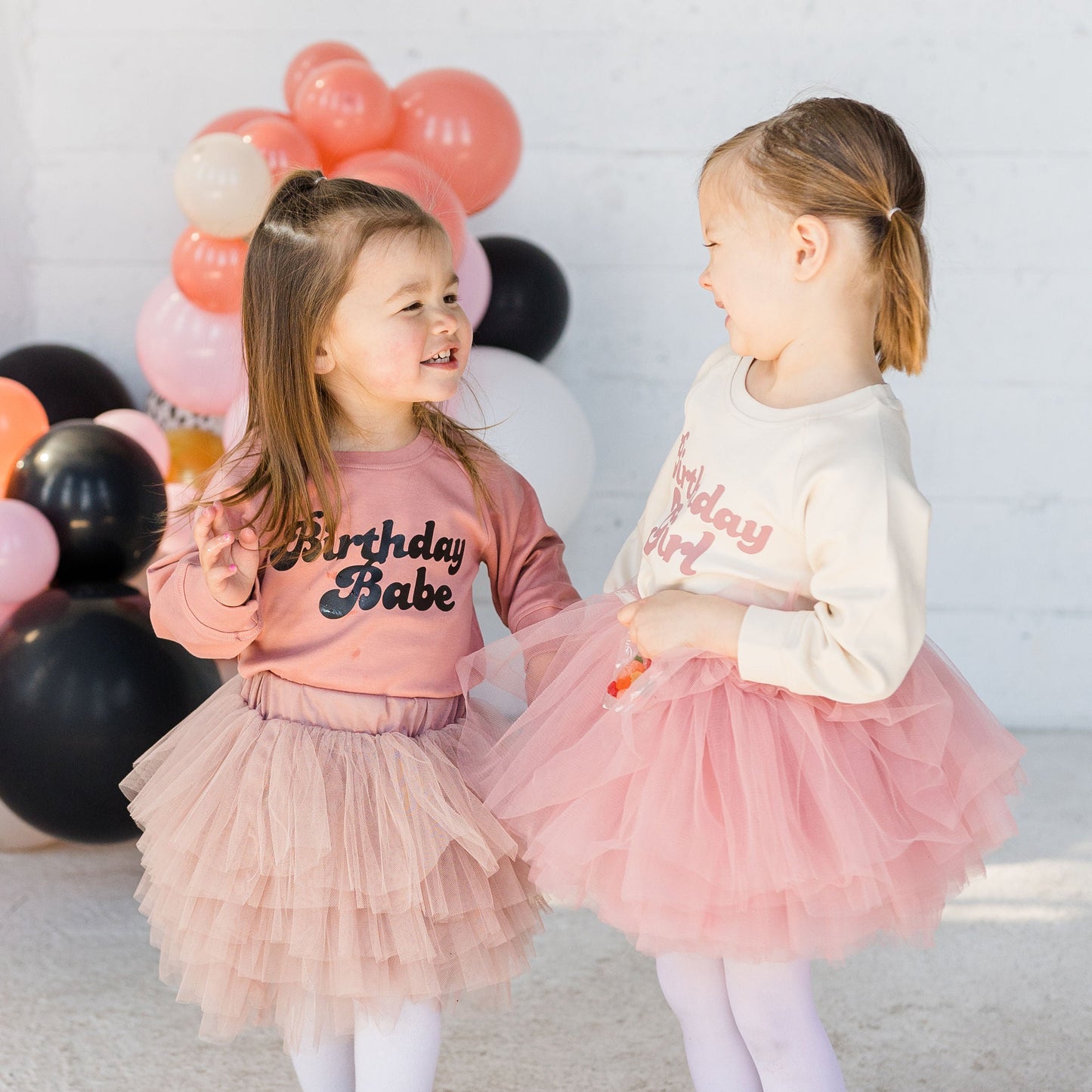 Two young girls in matching outfits with 'Birthday Babe' text, standing in front of balloons.