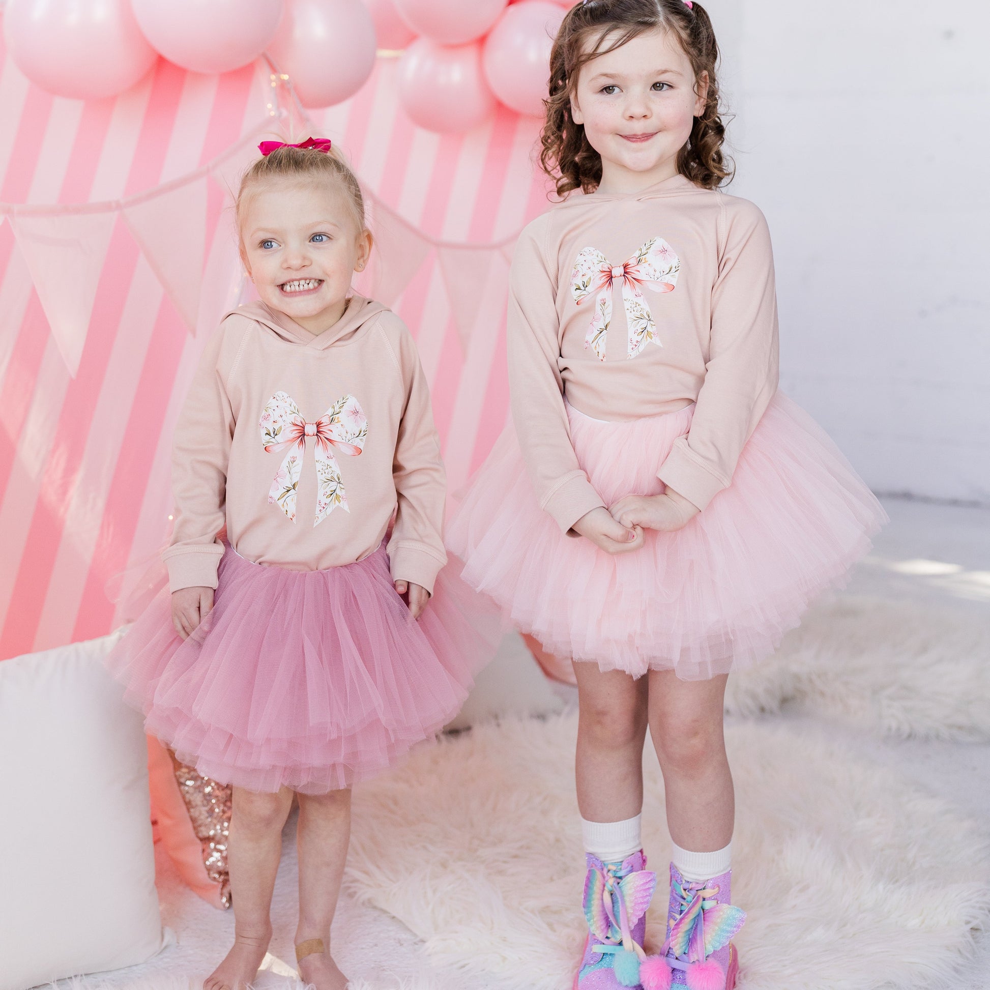 Two young girls in matching outfits with pink balloons and a pink arch in the background.
