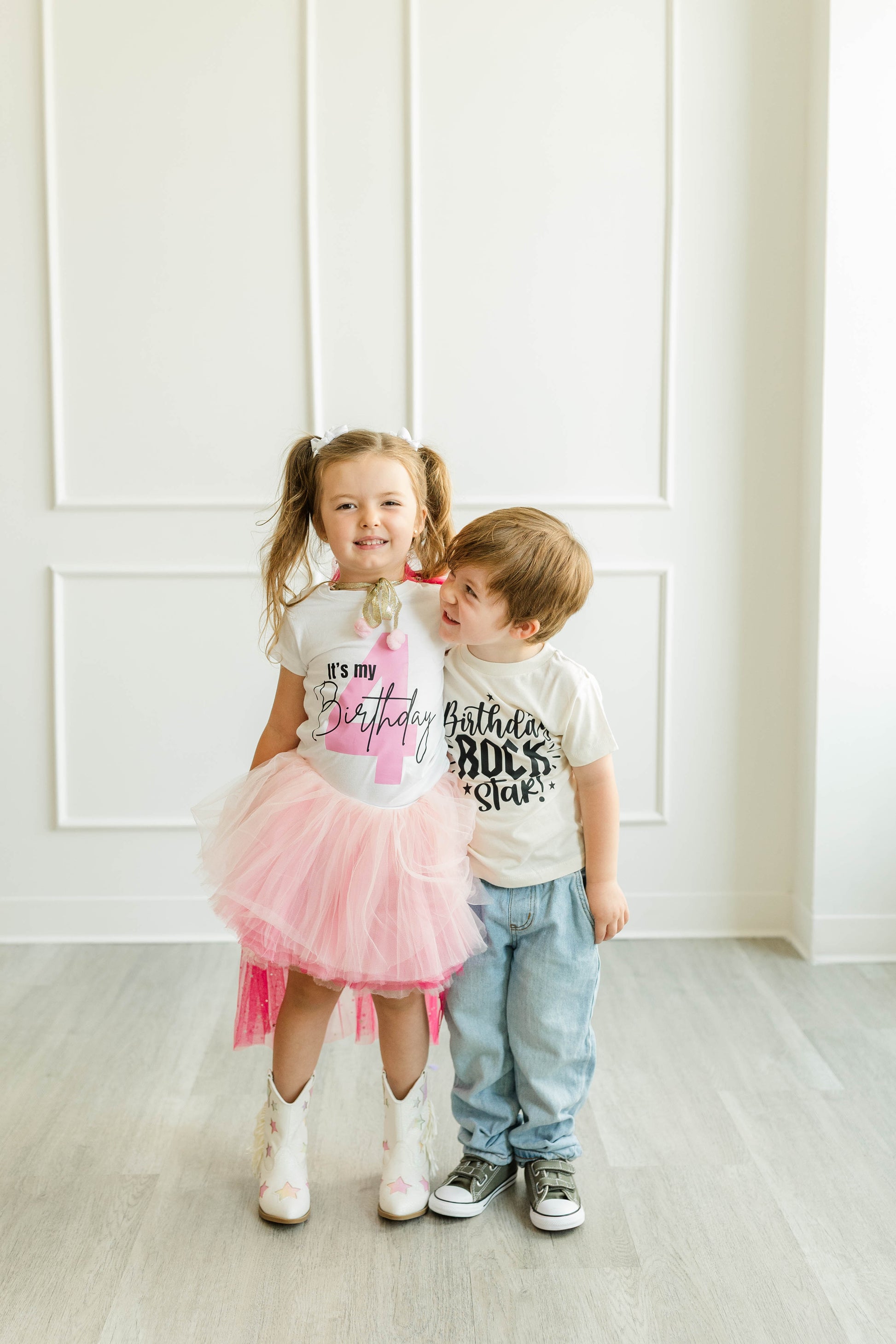 A young girl with pigtails and a boy standing together in a bright, white room. The girl wears a white t-shirt with 'it's my Birthday Rock Star' text and a pink tutu skirt, while the boy wears a matching white t-shirt with 'Birthday Rock Star' text and light blue jeans.
