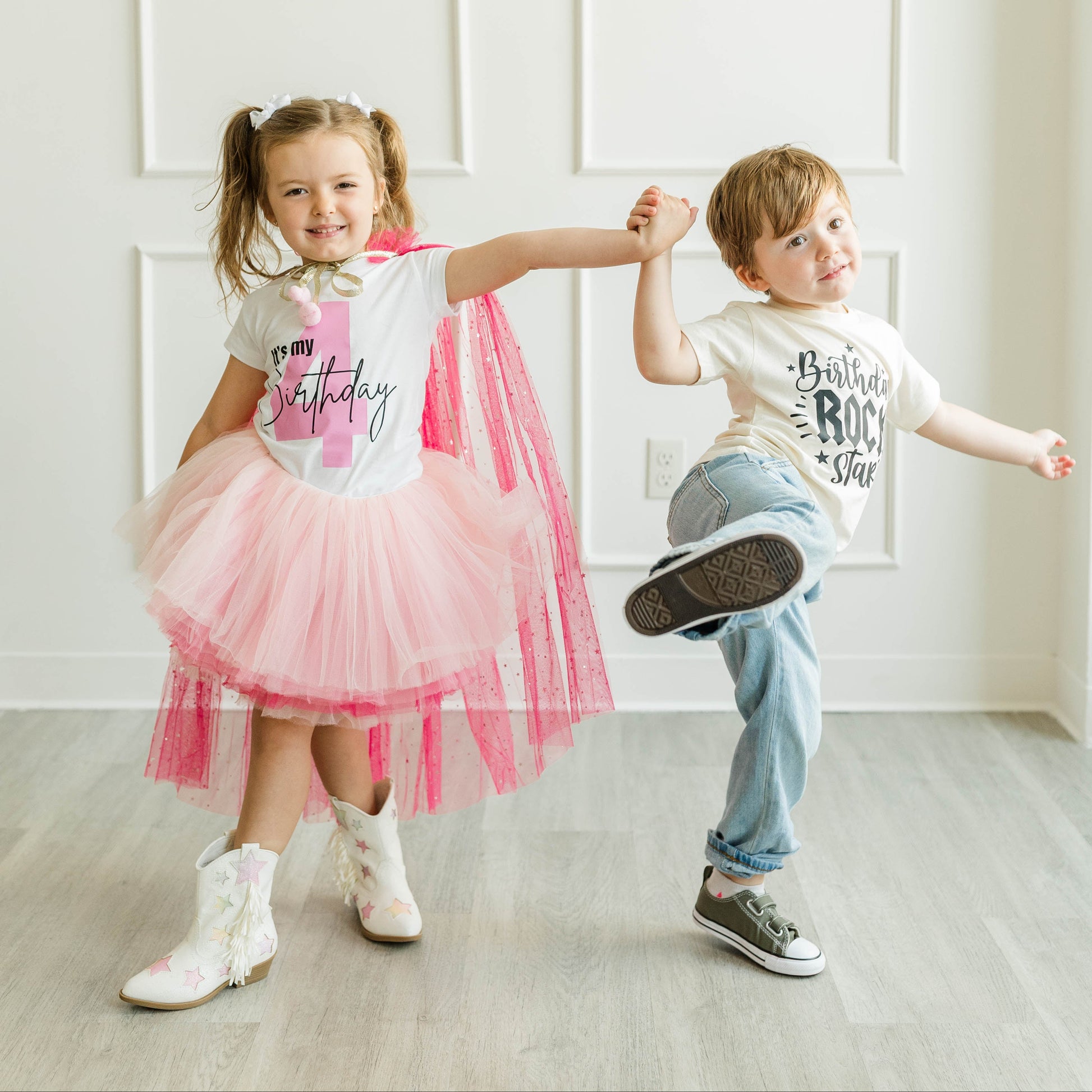 Two children dancing in a bright room. The girl wears a white 'BIRTHDAY' graphic tee with a pink tulle tutu and a pink cape, paired with white cowboy boots.