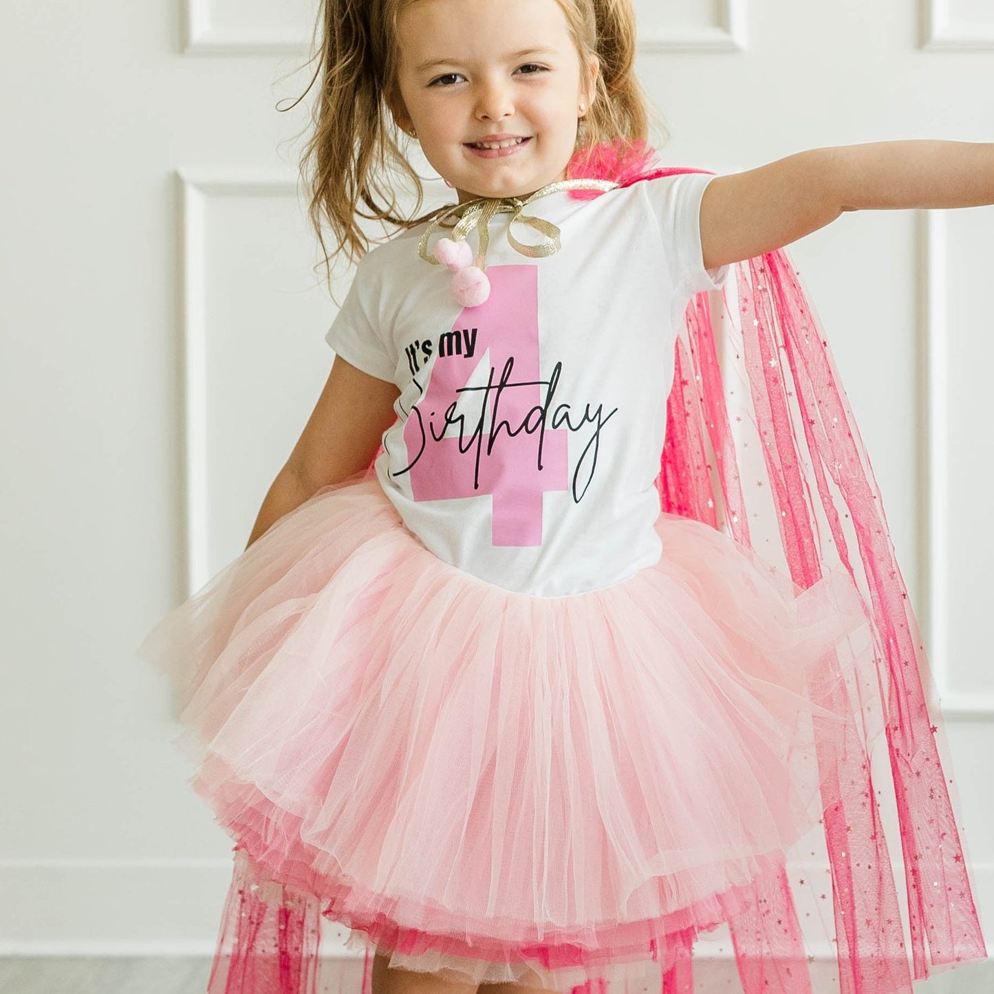 Two children dancing in a bright room. The girl wears a white 'BIRTHDAY' graphic tee with a pink tulle tutu and a pink cape, paired with white cowboy boots.