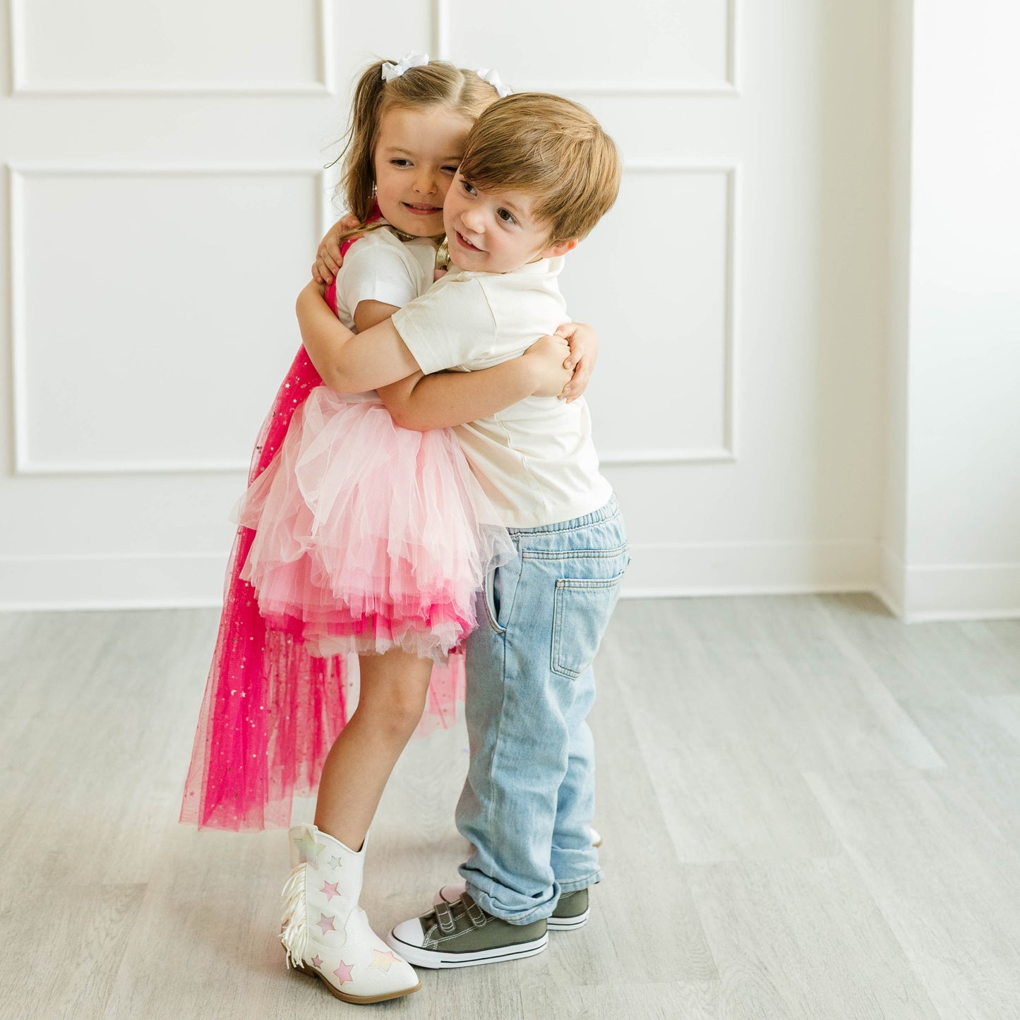 A young girl in a pink tulle tutu dress and white boots hugs a young boy wearing a white t-shirt, light blue jeans, and gray sneakers in a bright, modern room with white paneled walls and light wood floors.
