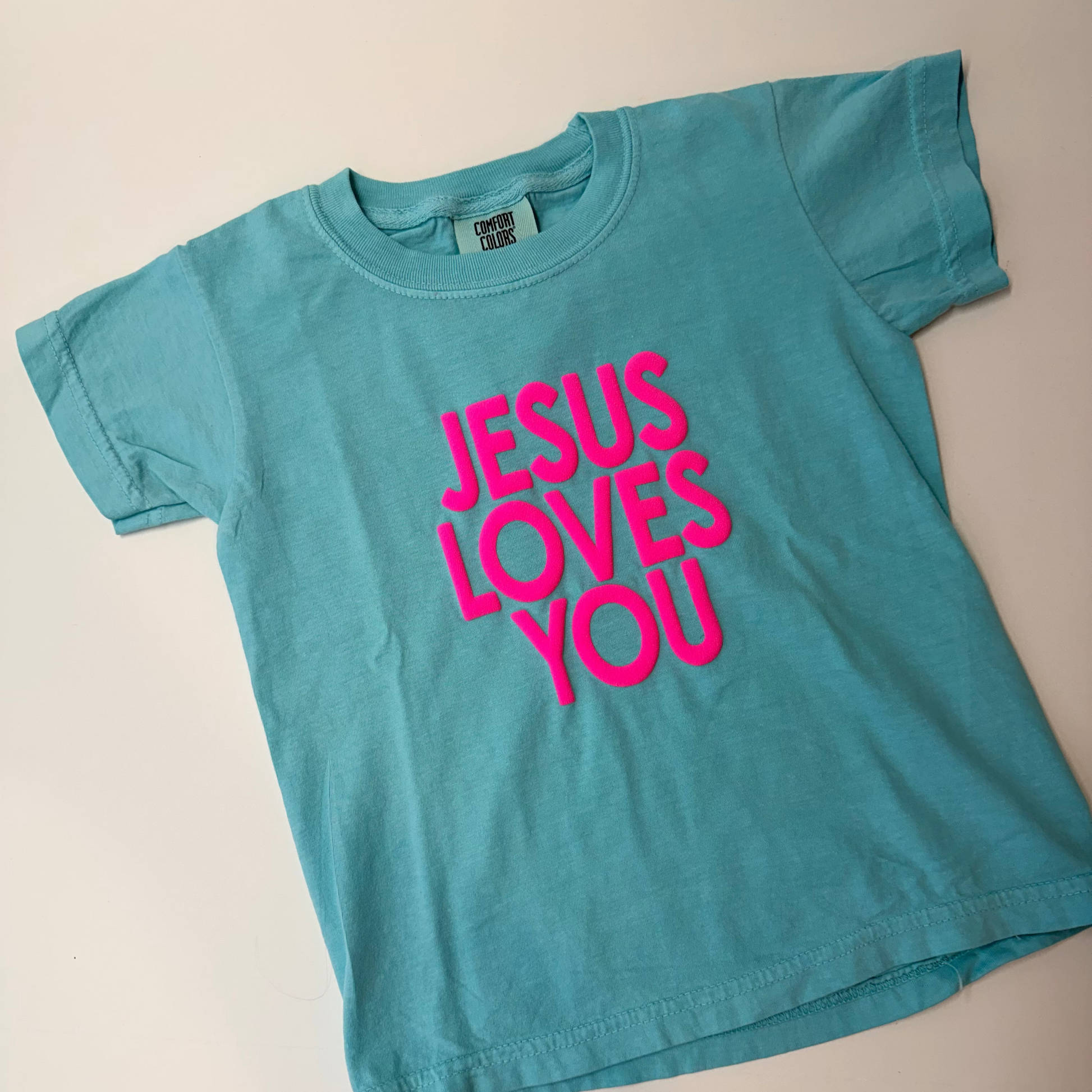 Blue t-shirt with 'Jesus Loves You' in pink on a beige background