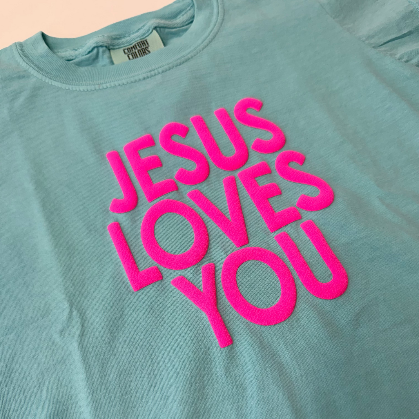 Light blue t-shirt with 'Jesus Loves You' in pink text on a beige background