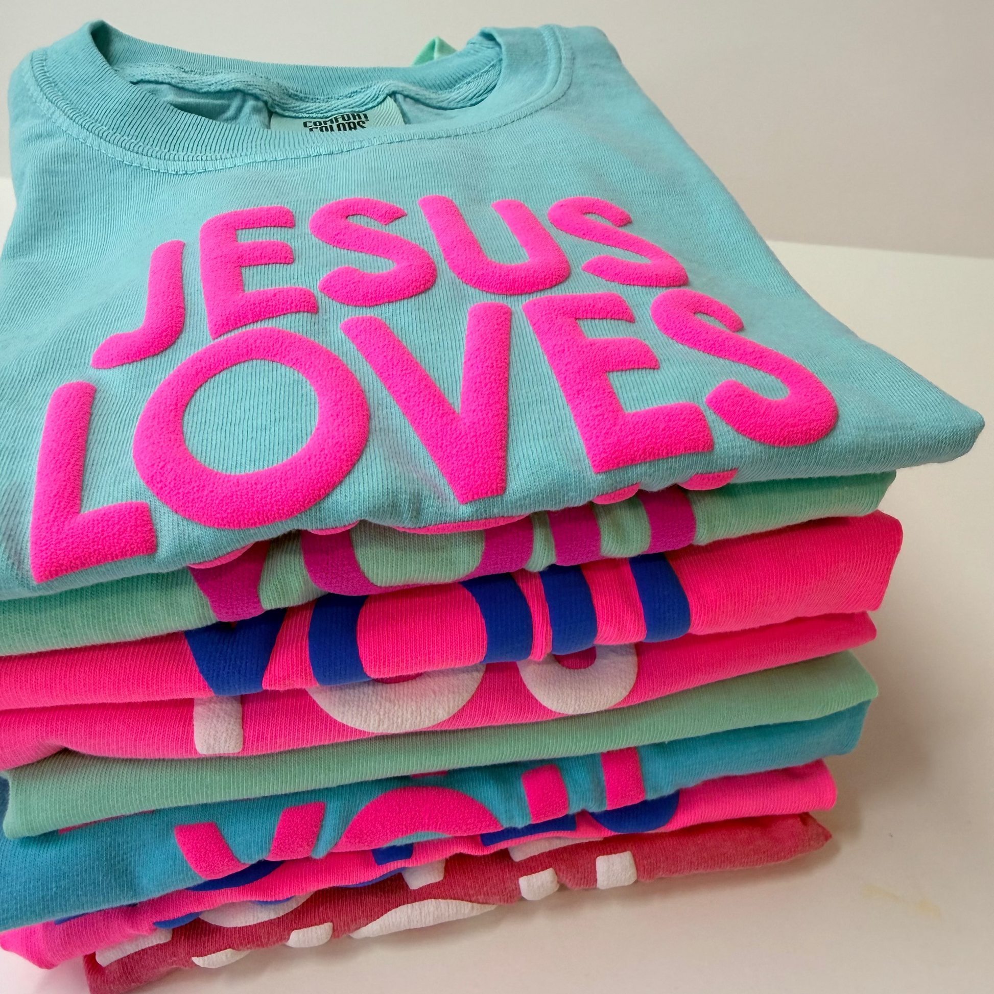 Stack of colorful t-shirts with 'Jesus Loves' text on a white surface