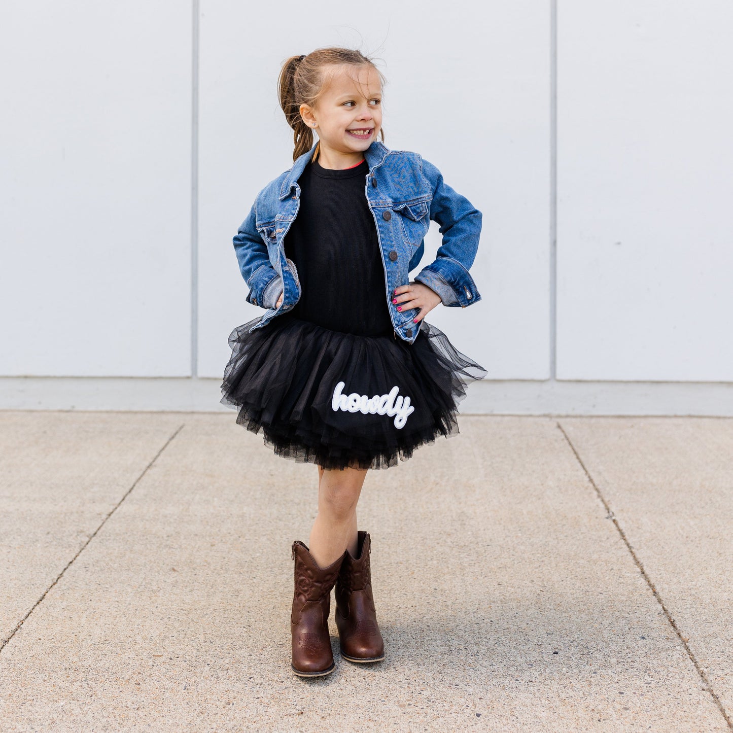 Child wearing a denim jacket, black dress with 'happy' text, and brown boots against a white wall.