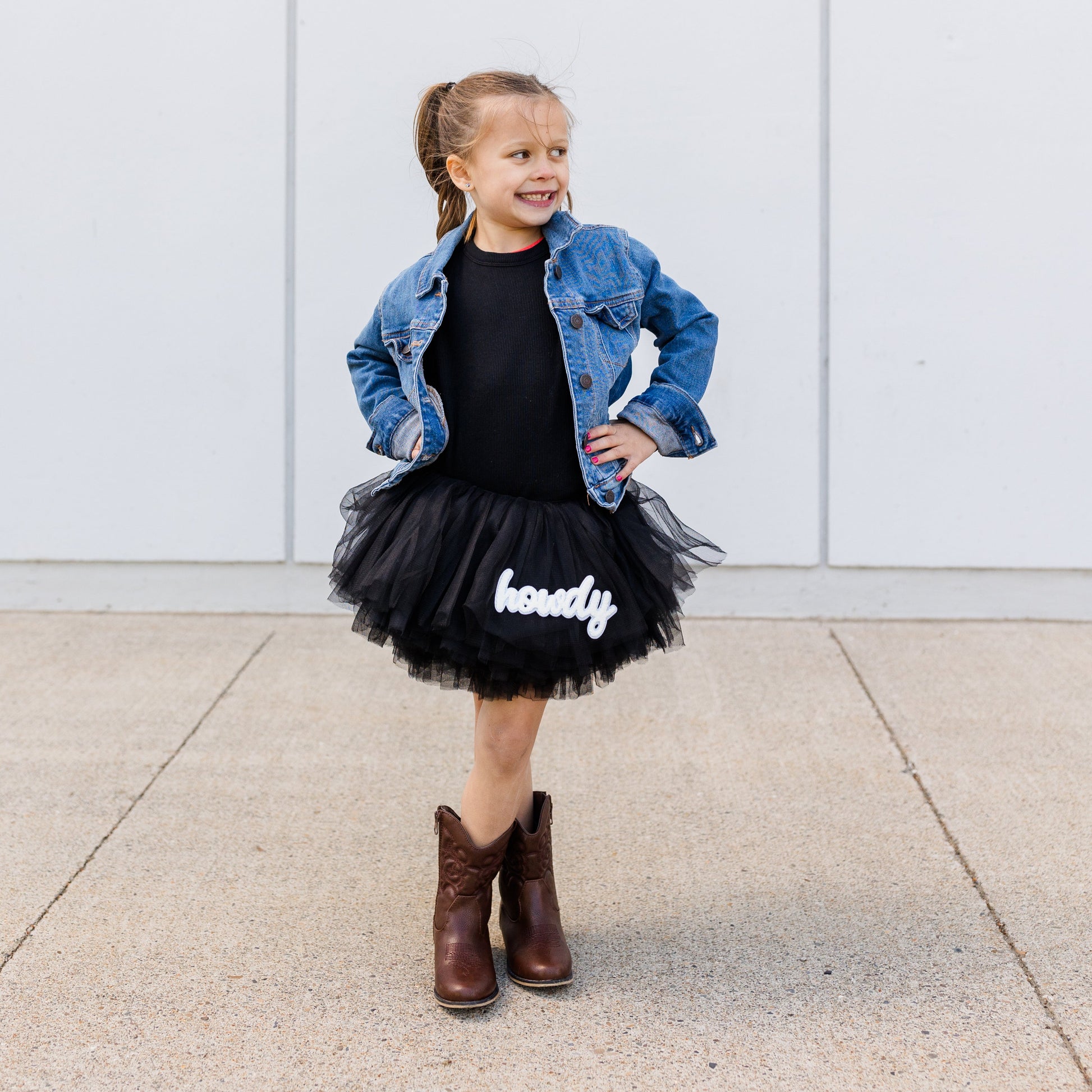Child wearing a denim jacket, black dress with 'happy' text, and brown boots against a white wall.