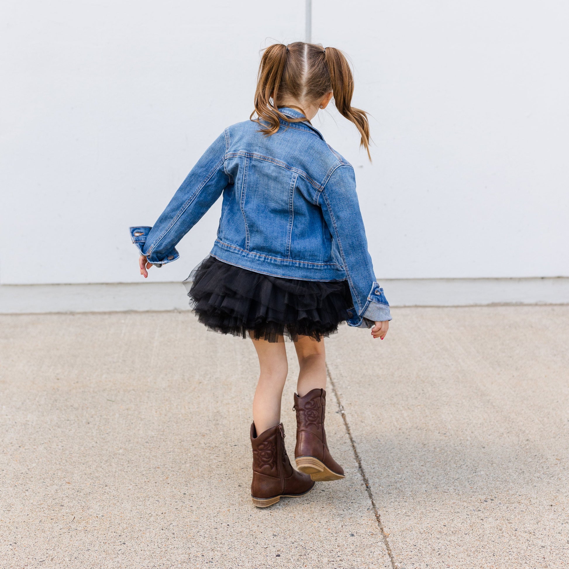 Child wearing a denim jacket and black skirt with brown boots against a white wall.