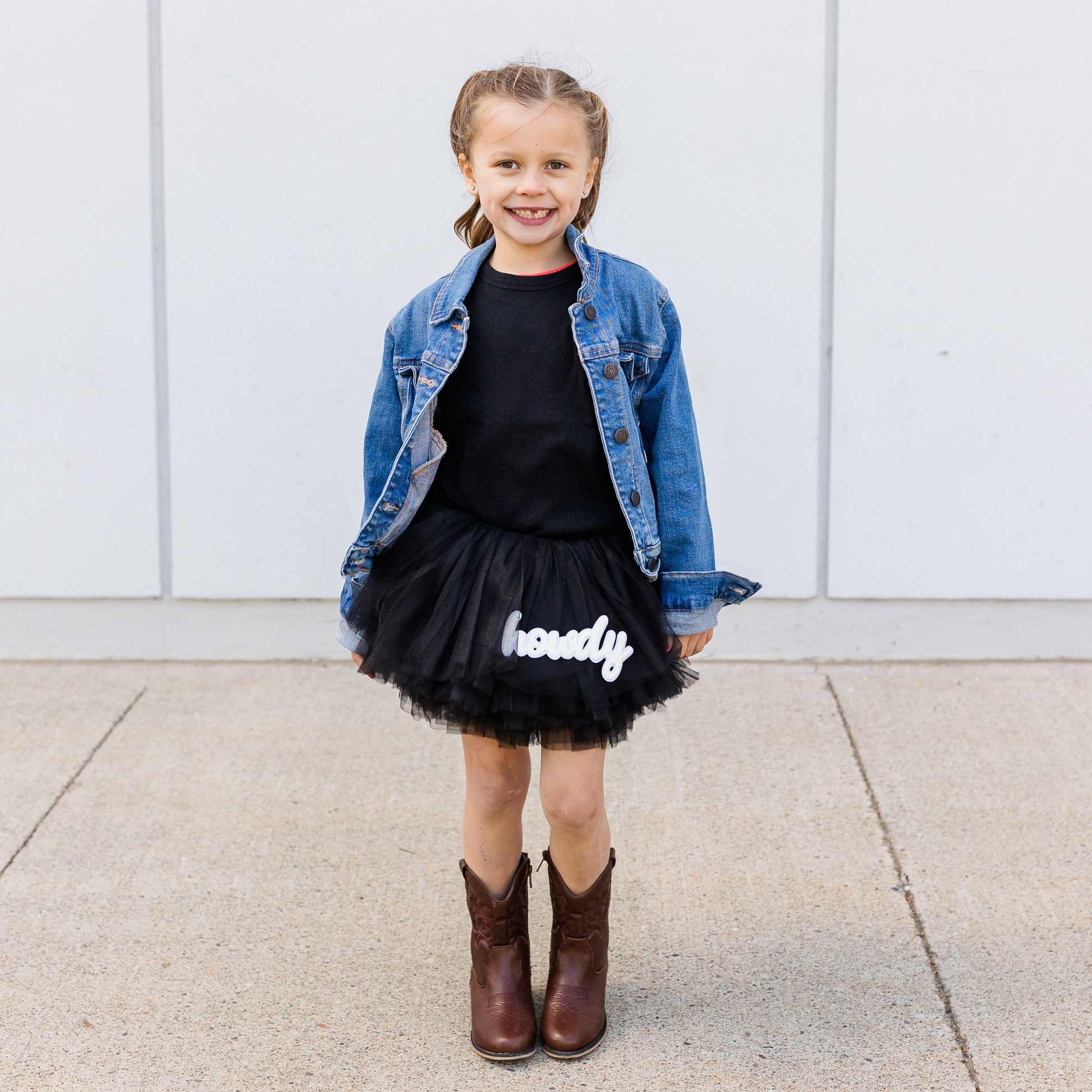 Young girl wearing a denim jacket, black shirt, tutu skirt with ‘howdy" text, and brown boots against a white wall.