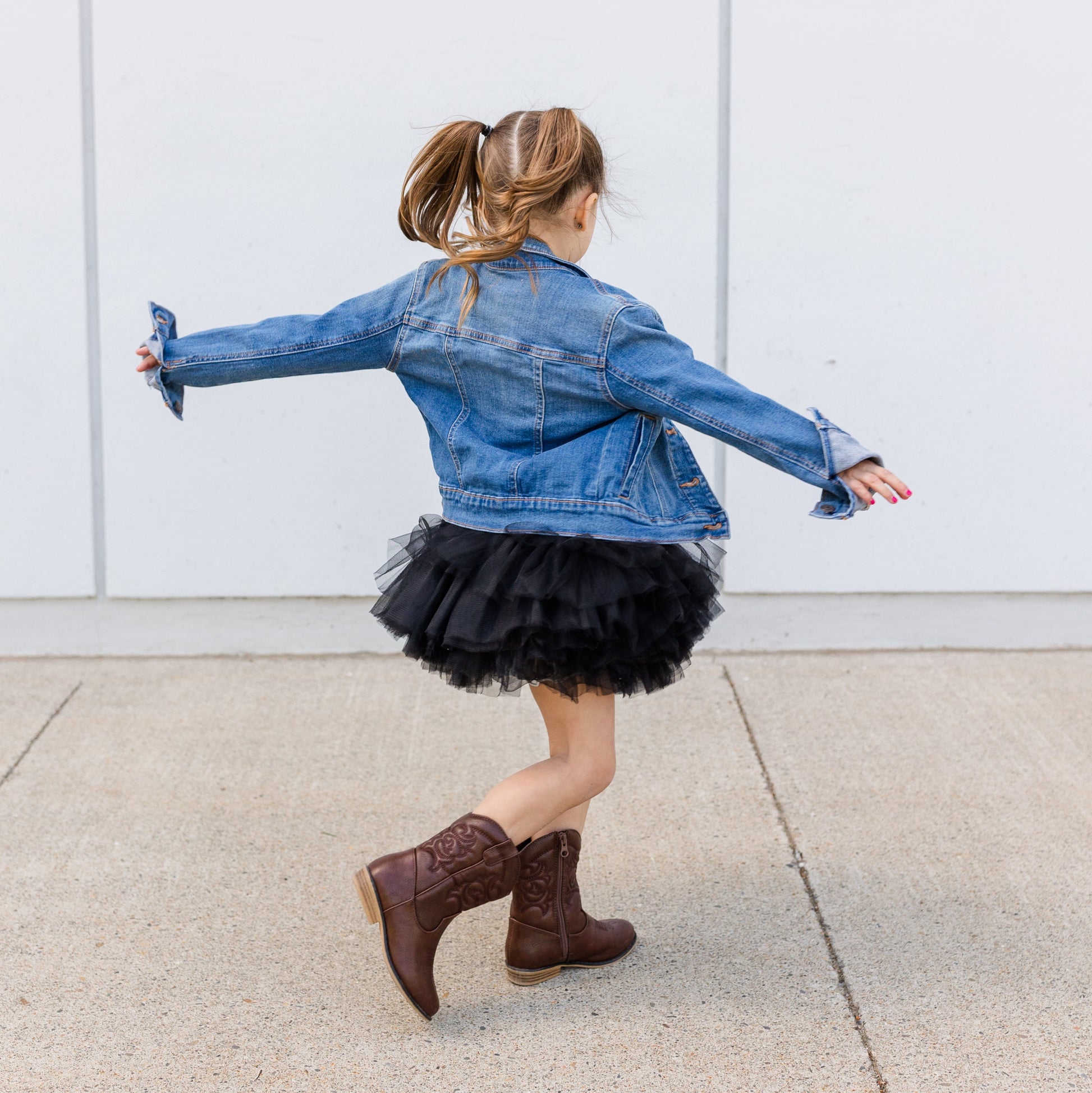 Person wearing a denim jacket and black tutu skirt with brown boots, standing against a white wall.