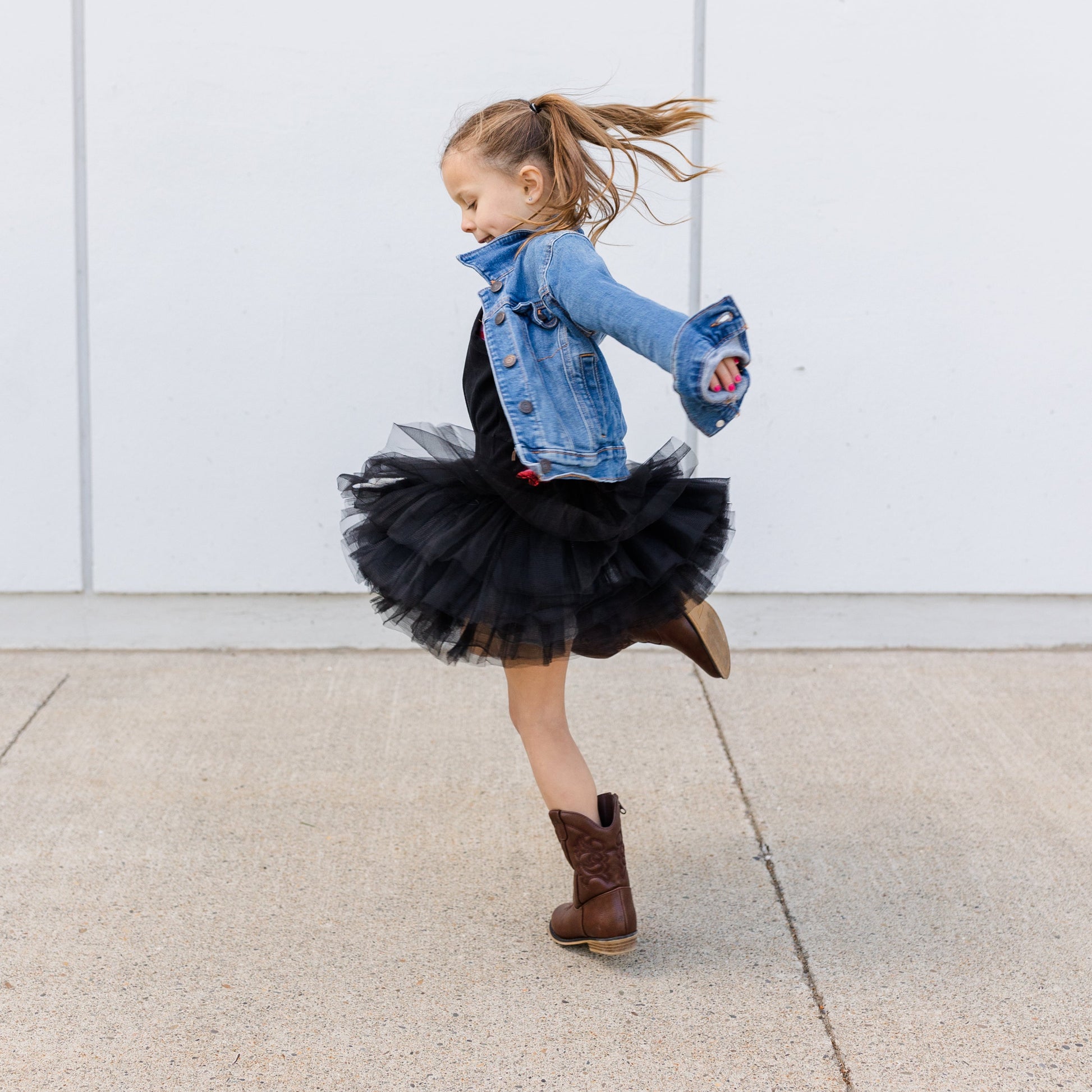 Child in denim jacket and black tutu dancing against a white wall.