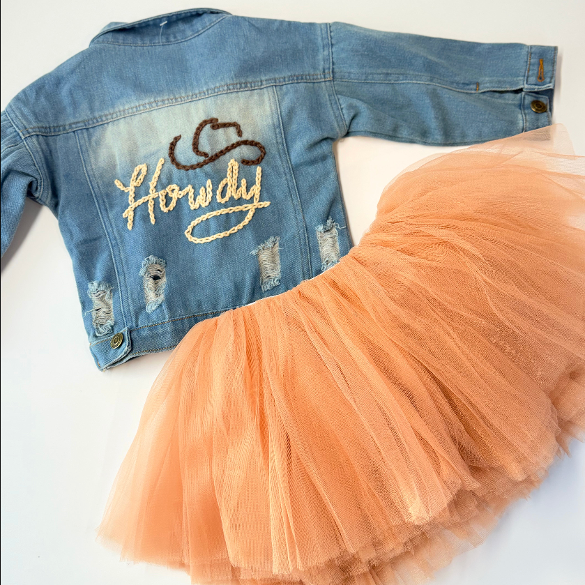 Toddler distressed jean jacket with Howdy in tan stitching and darker brown stitching cowboy hat, hand stitched. Paired with The Glam Goat caramel tutu that is not included with the jacket. Laid on a white background 