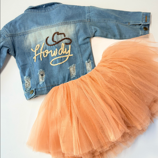 Toddler distressed jean jacket with Howdy in tan stitching and darker brown stitching cowboy hat, hand stitched. Paired with The Glam Goat caramel tutu that is not included with the jacket. Laid on a white background 