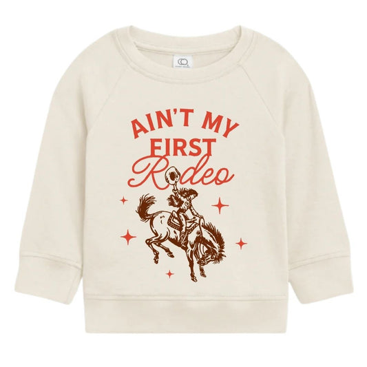 Cream organic cotton kids long sleeve sweatshirt with red “Ain’t My First Rodeo” graphic and cowboy riding bucking horse.
