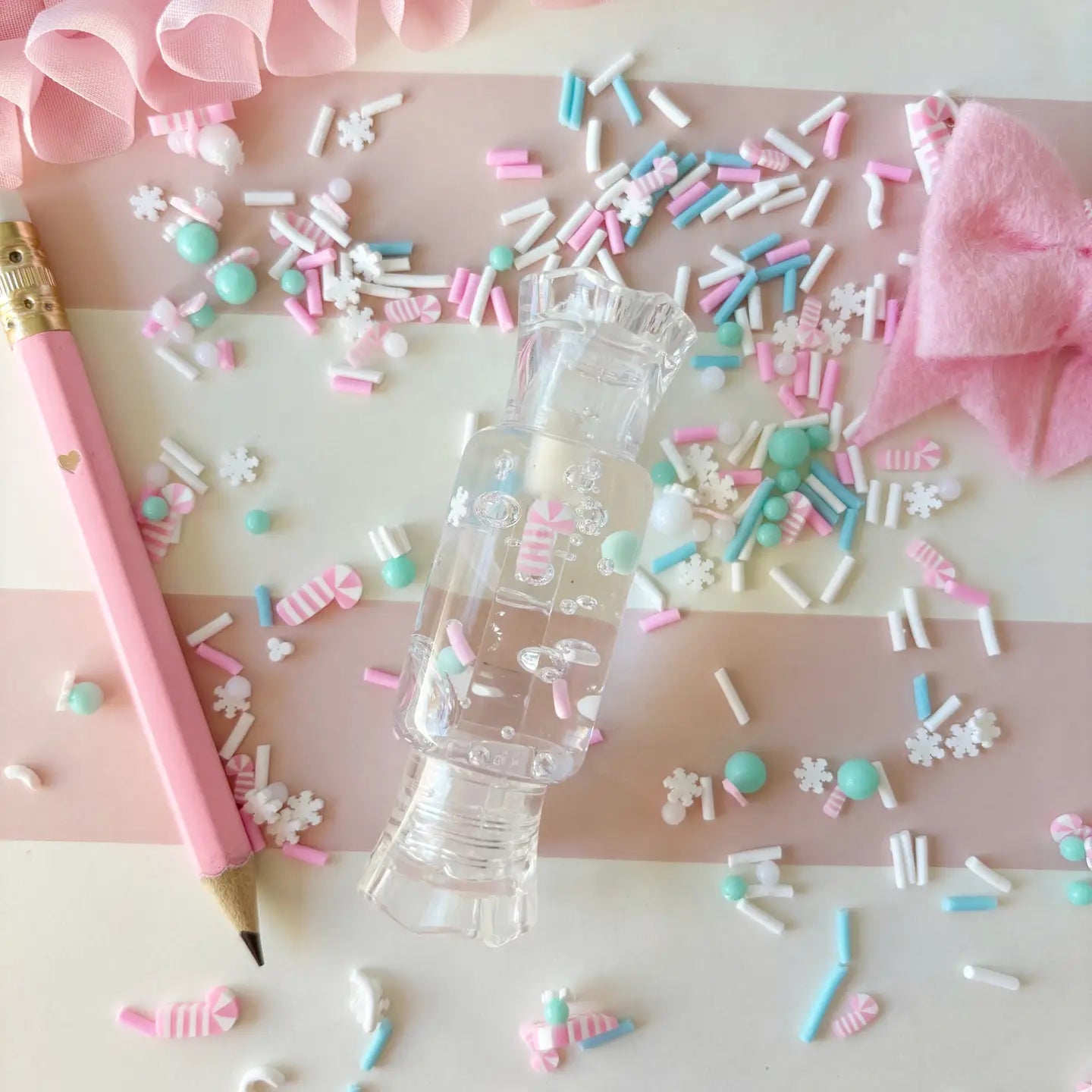 Clear plastic bottle with pink cap on a pink and white striped background with colorful sprinkles and a pink pencil.
