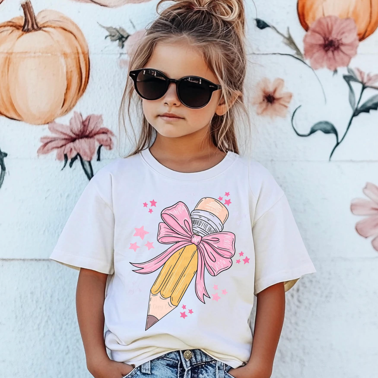 Young girl wearing sunglasses and a white t-shirt with a large yellow pencil wrapped in a pink bow and surrounded by pink stars, standing in front of a mural with pumpkins and flowers.
